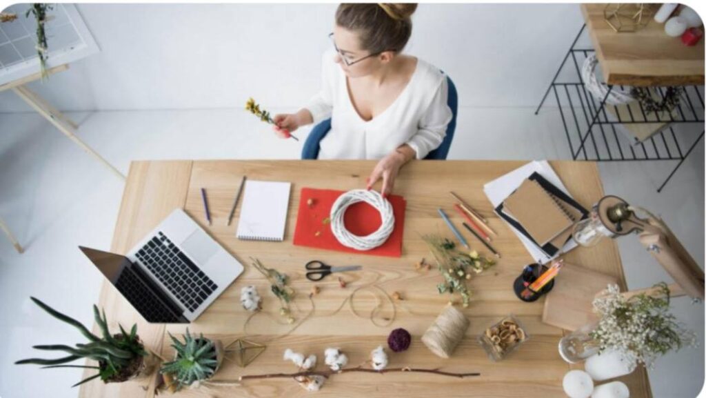 Mulher criando peças de artesanato digital em mesa de madeira com notebook e materiais sustentáveis, representando e-commerce verde e artesanato digital.